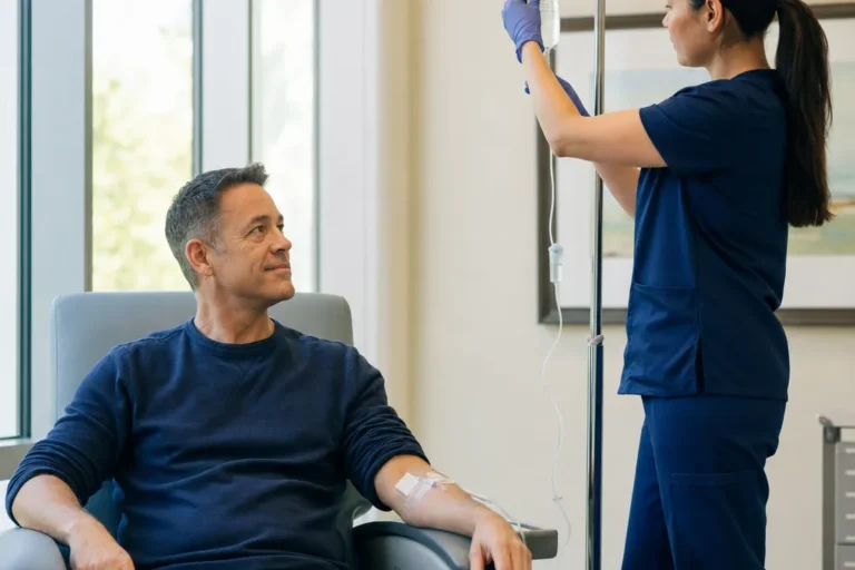 Adult patient resting in a treatment chair with an IV in his forearm while a clinician checks the fluid bag