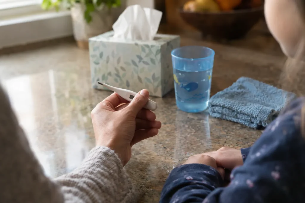 Parent's hand holding a thermometer beside tissues and a child's cup