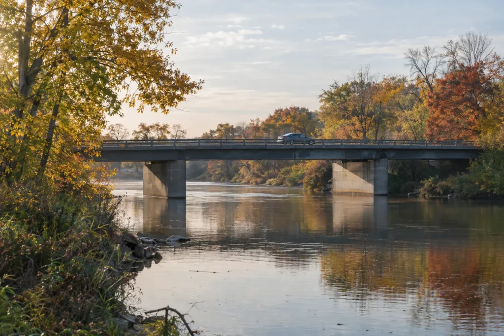 A car crosses a road bridge over a tree-lined river in soft morning light